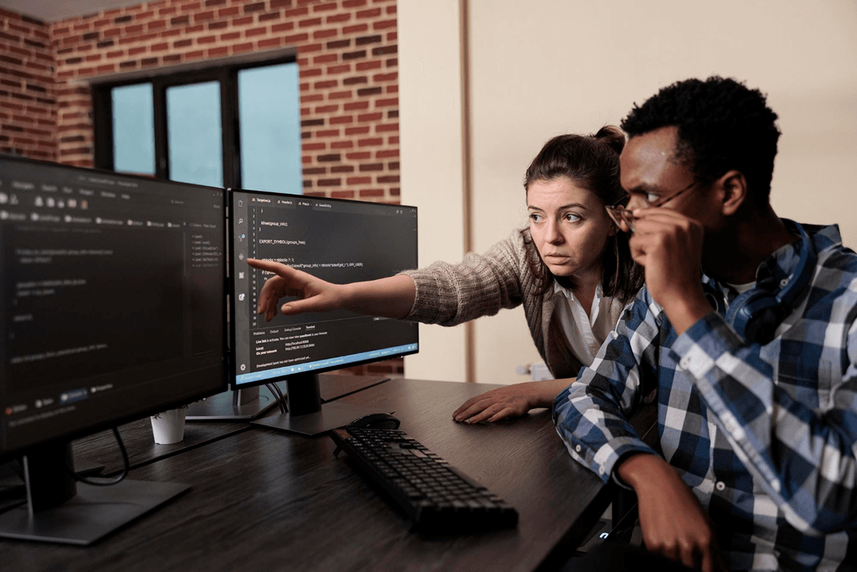 Two developers working together.  A woman is pointing at code displayed on a computer monitor, guiding her male colleague.