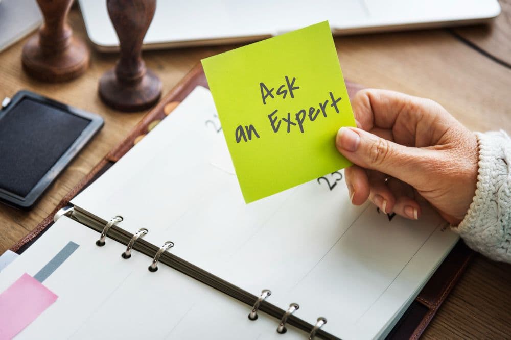Open Planner With a Person's Hand Holding a Sticky Note That Says 'Ask an Expert'.