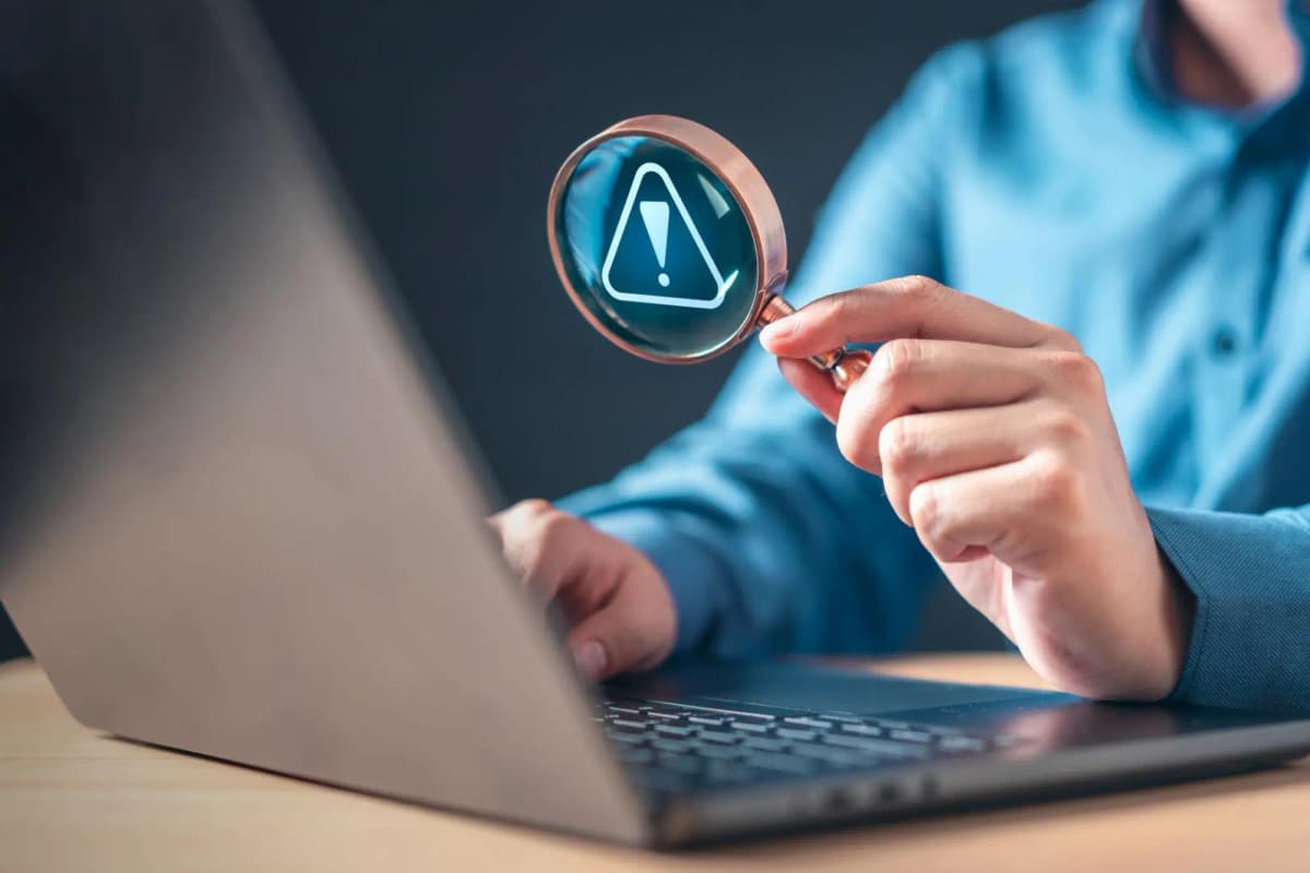 A person uses a magnifying glass over a laptop screen highlighting a warning symbol, representing identifying digital issues.