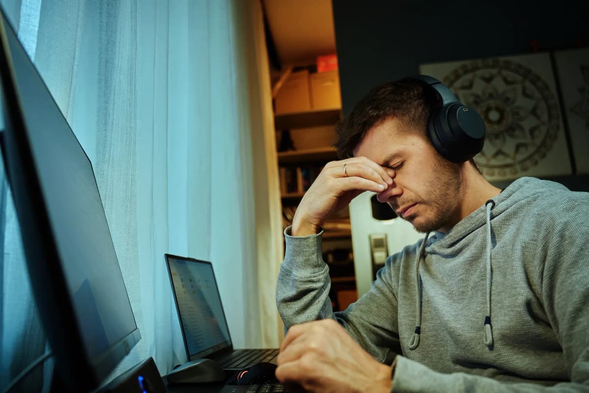 Man wearing headphones rubbing his eyes while working on a laptop, showing frustration during digital tasks