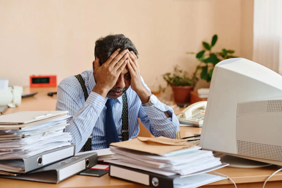 A man sits at a cluttered desk with stacks of documents, holding his head, showing stress from overwhelming workload.