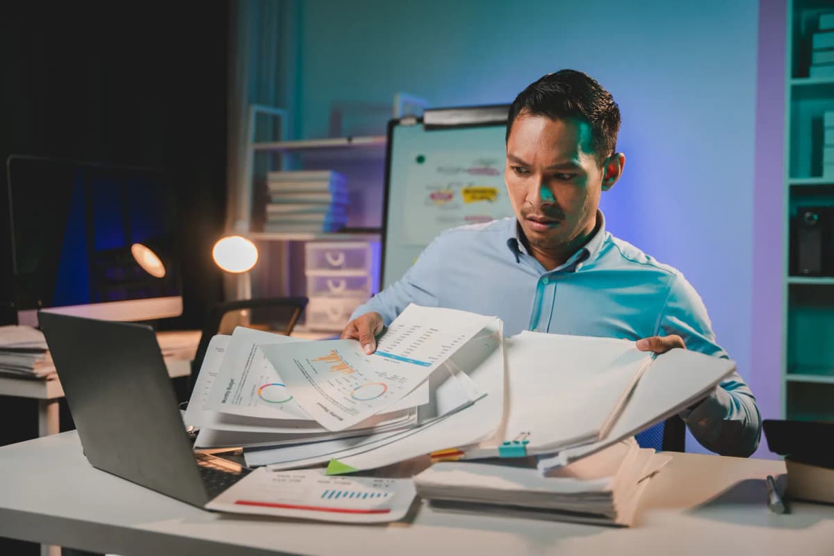 A male office worker reviewing large stacks of documents at a desk, appearing overwhelmed by paperwork
