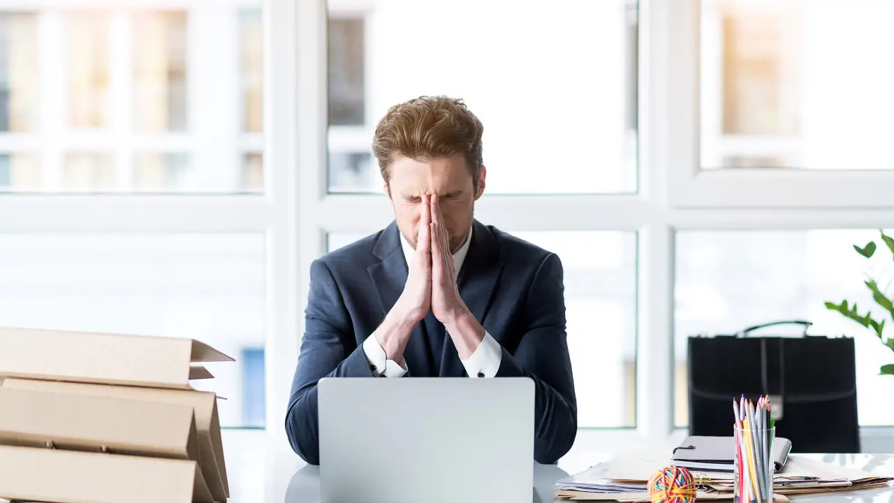 Office worker sitting at a desk with hands pressed together in stress, surrounded by paperwork and a laptop, representing workload pressure and limited capacity