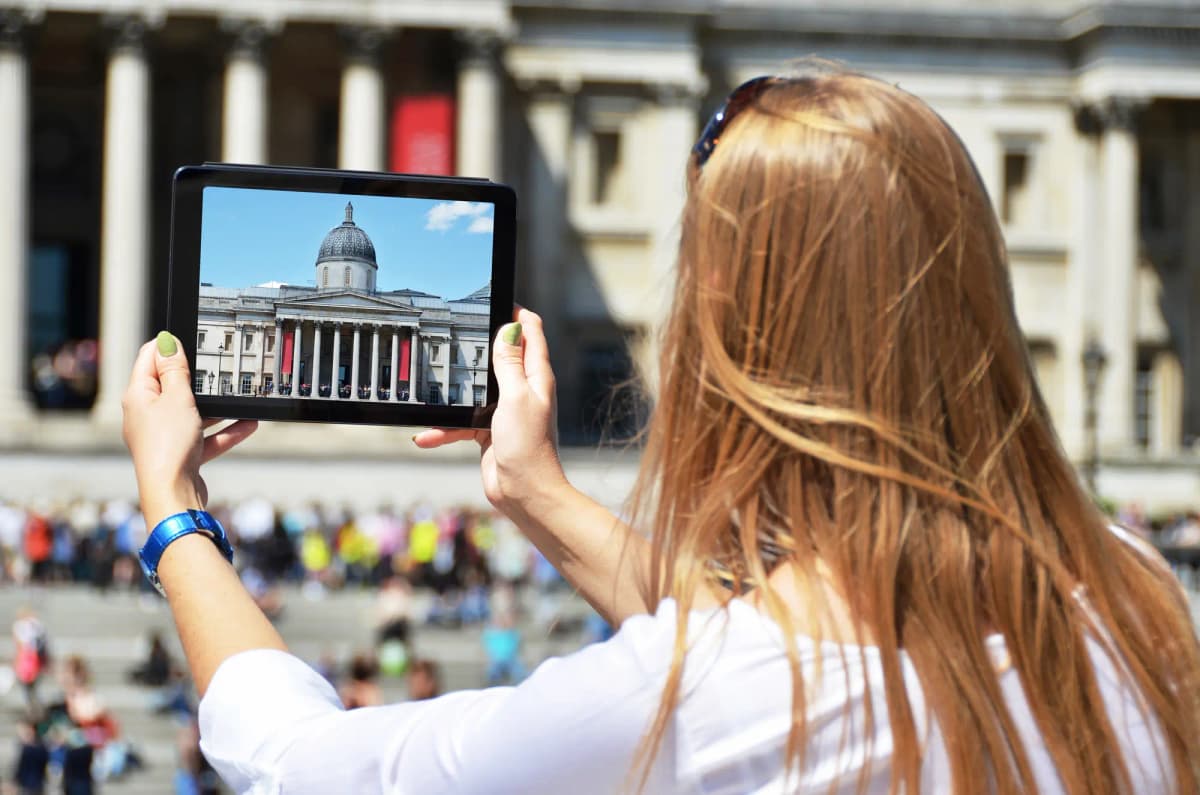 A blonde woman holding a tablet capturing a government building with crowd, showing publicly shared civic video content requiring accessibility.