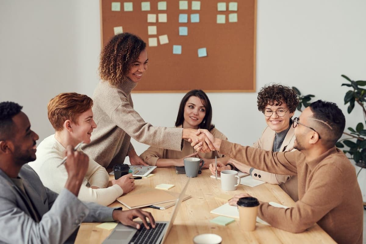 Six people in a meeting room. A woman is shaking hands with a man while others smile and observe.