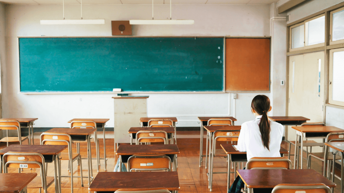 Student sitting alone in a nearly empty classroom facing the chalkboard, with rows of empty desks in front, symbolizing educational isolation.