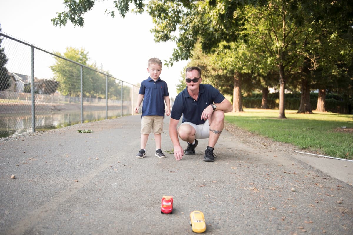 A blind father sits on the ground in a park, playing with toy cars with his young son, who is standing beside him. This moment showcases a blind parent's love and engagement, challenging misconceptions.