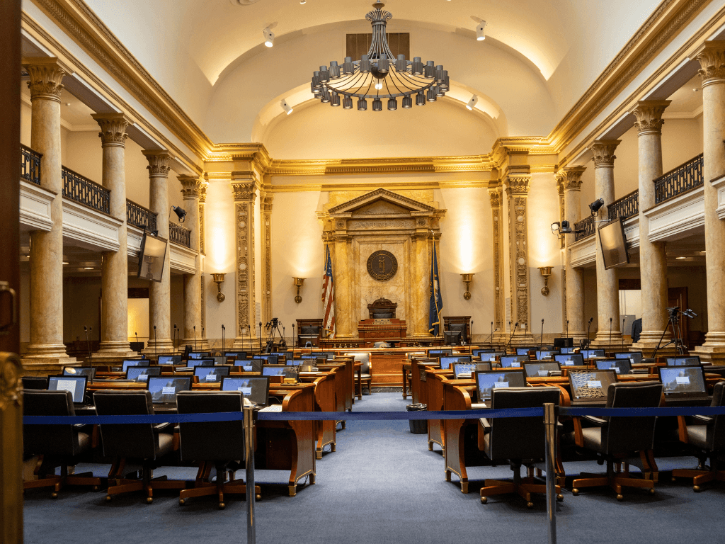 Interior of a formal government chamber with rows of desks, computer monitors, and American flags, viewed from the entrance.