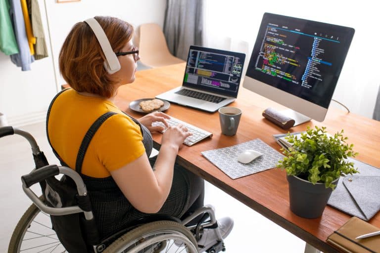 Female freelance programmer in modern headphones sitting in wheelchair and using computers while coding.