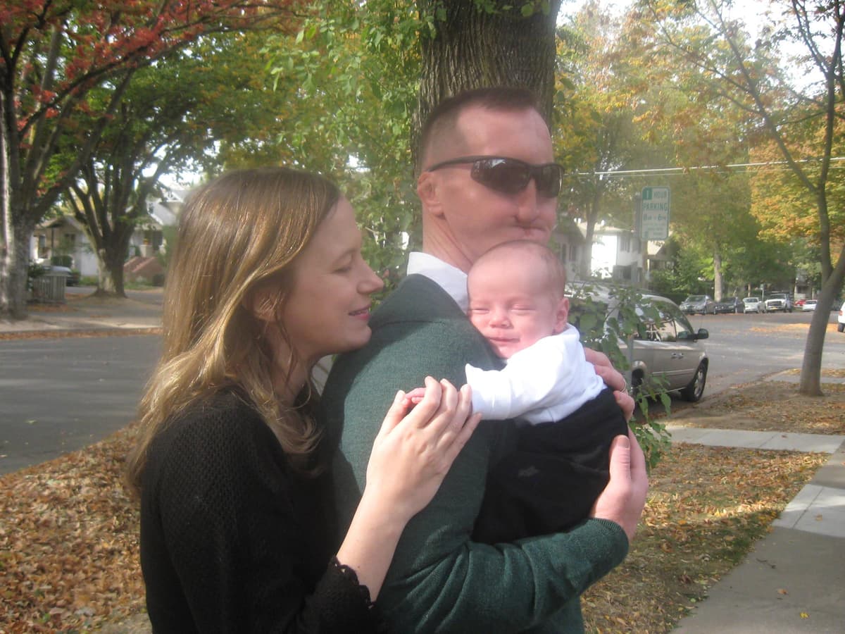 A blind father holds his sleeping baby, who appears to be smiling, while the mother stands behind them, smiling warmly.