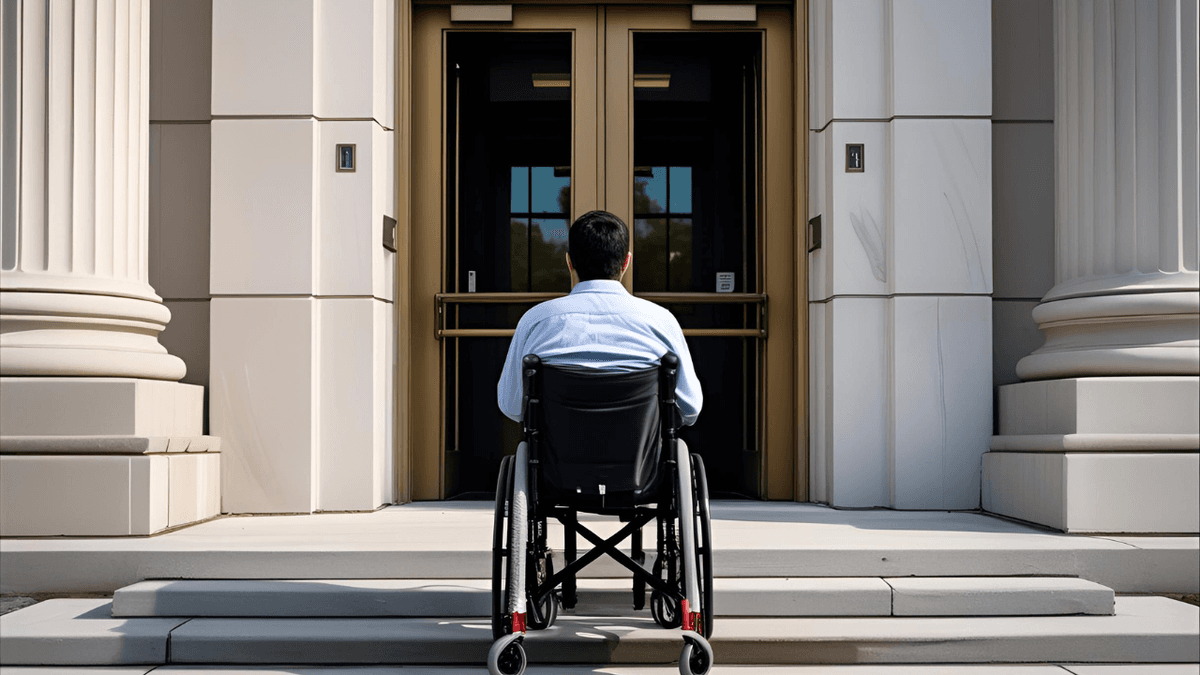 A man using a wheelchair facing the steps of a government building without a ramp, symbolizing barriers to accessibility compliance.