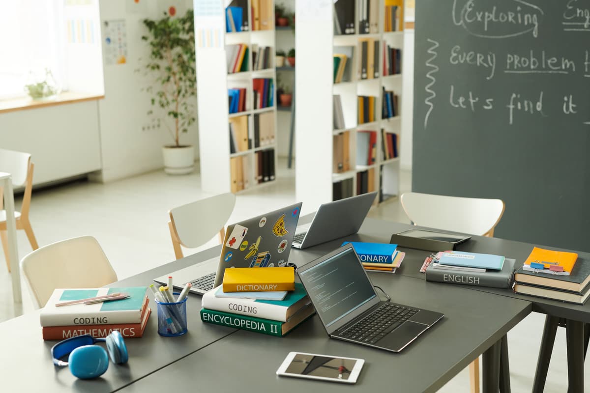 On top of a classroom table are laptops, a tablet with headphones, along with an English and coding text books, illustrating the importance of digital accessibility and Section 504 in K-12 schools