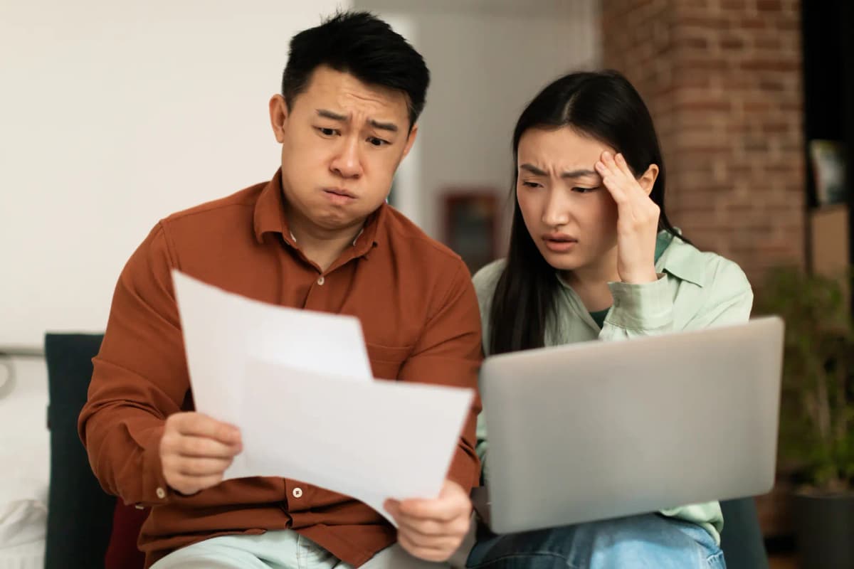 Two people sitting side by side looking worried while reviewing a document. The man holds a sheet of paper with a concerned expression, while the woman holds her head and looks at a laptop beside her, suggesting confusion or stress over the information.