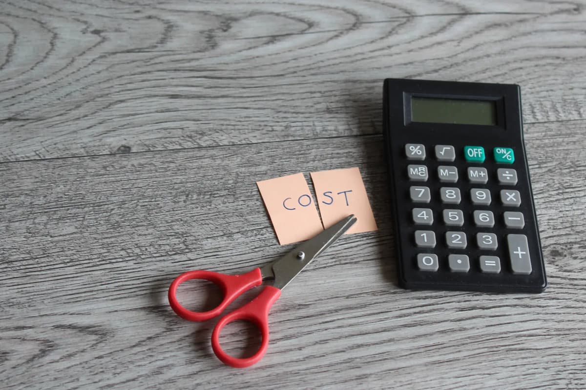 Red scissors cutting note labeled cost beside black calculator on a wooden surface