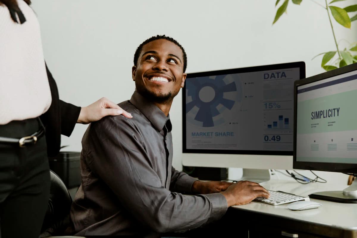 A male office worker at his computer desk, receiving guidance from a colleague, he looks at her smiling, while she stands beside him and holds his shoulders