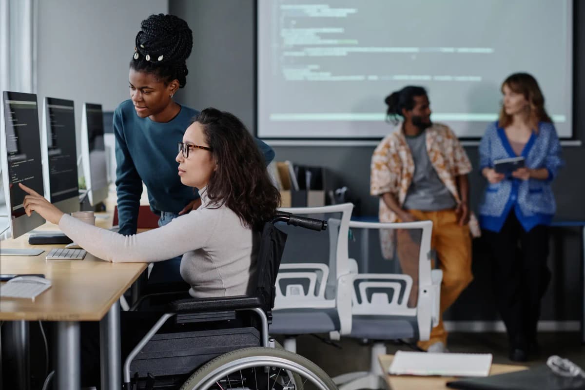A diverse group of people in a technology classroom collaborate around computer screens. A woman using a wheelchair works at a computer while others discuss coding and digital accessibility in the background.