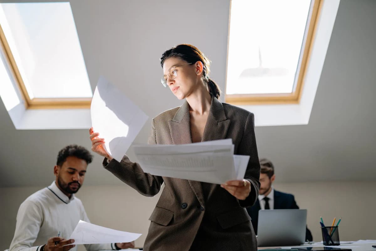 Woman standing in an office reviewing printed documents while colleagues work at a desk with a laptop