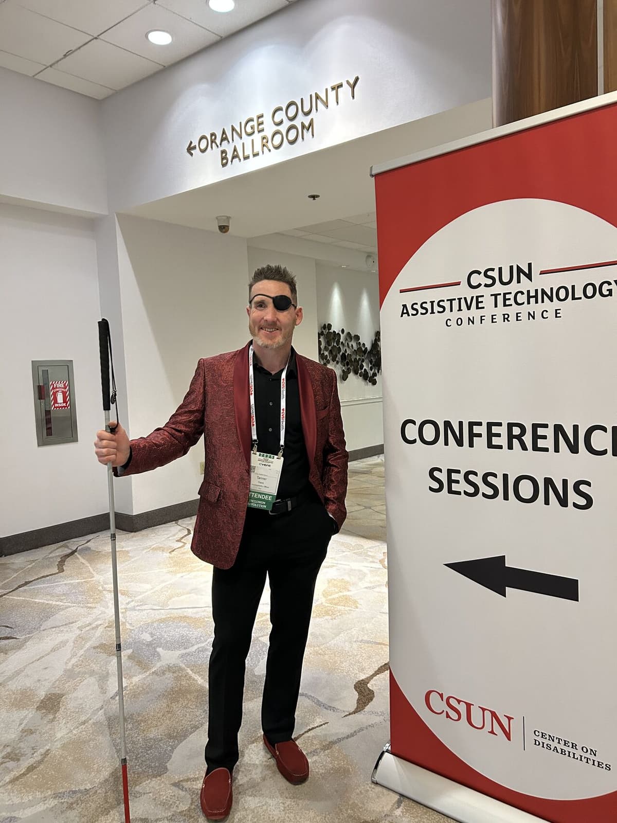 Tanner Gers, CEO of AccessAbility Officer and DARTSuite at CSUN Assistive Technology Conference 2025 (He stands confidently and gratefully wearing his black eyepatch, fiery red coat, his cane, black shirt and pants)