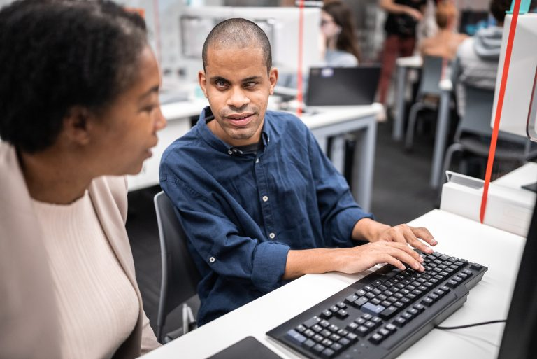 A blind man uses a keyboard in a computer lab while speaking with a colleague, illustrating accessibility and inclusion in the workplace.