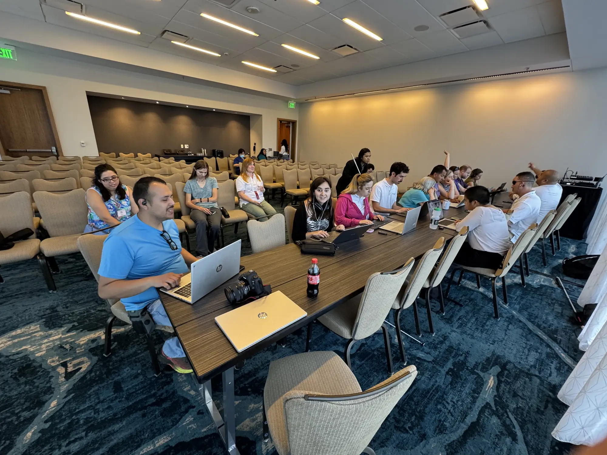 Testathon participants and AO Team sit along a long conference table in a meeting room, working on laptops. Some wear headphones or earpieces, and a white cane is visible at the right edge.
