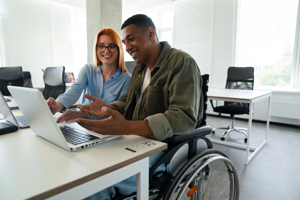 Two people in a workplace. The man, who uses a wheelchair, is working on a computer.