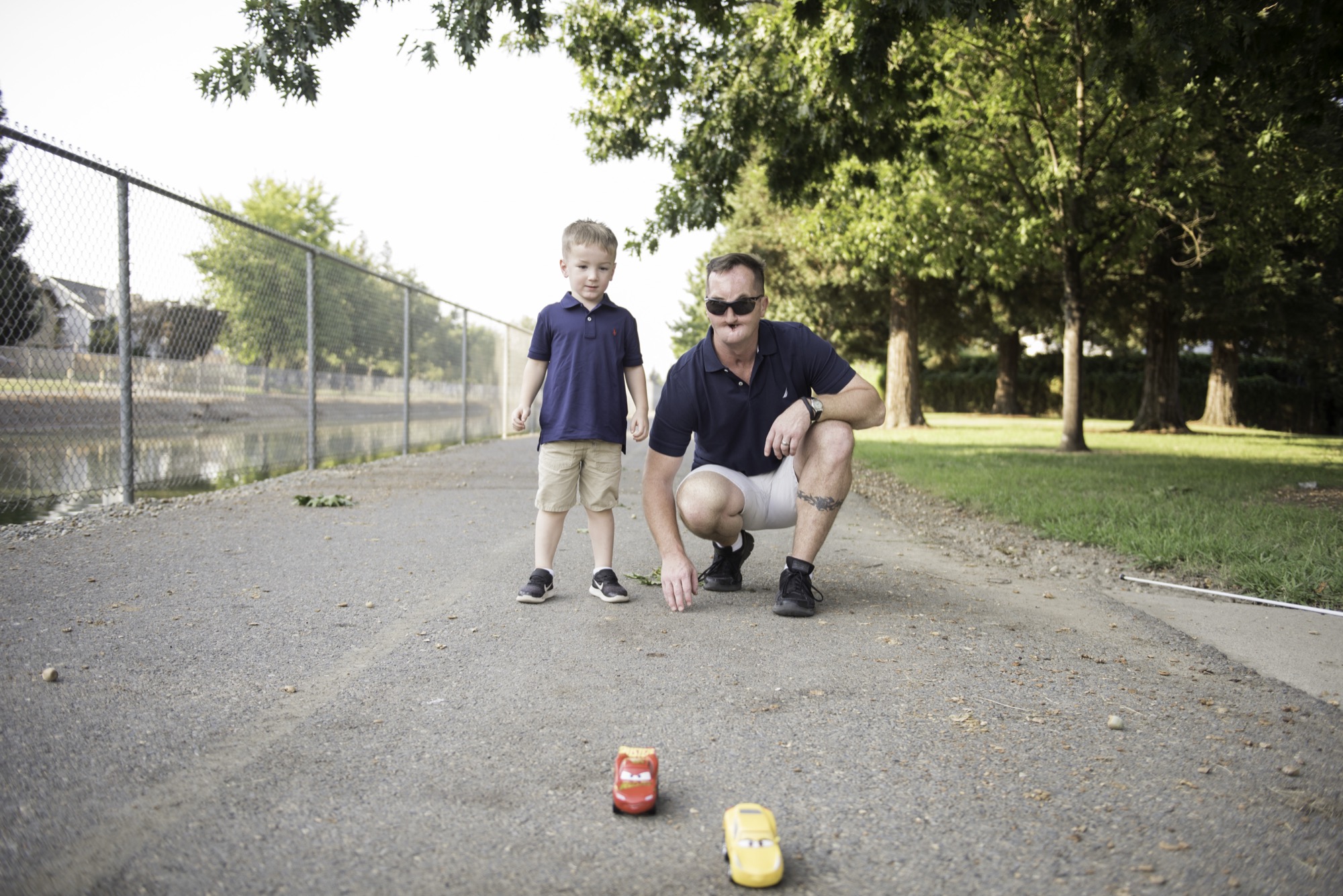 A blind father sits on the ground in a park, playing with toy cars with his young son, who is standing beside him. This moment showcases a blind parent's love and engagement, challenging misconceptions.