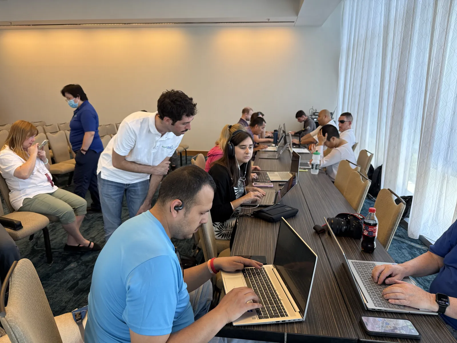 Participants at an AccessAbility Officer testathon during the National Federation of the Blind national convention, working at laptops along a conference table. Several are using headsets and earpieces for screen readers. Testing Manager Liam Colburn observes over a participant's shoulder.