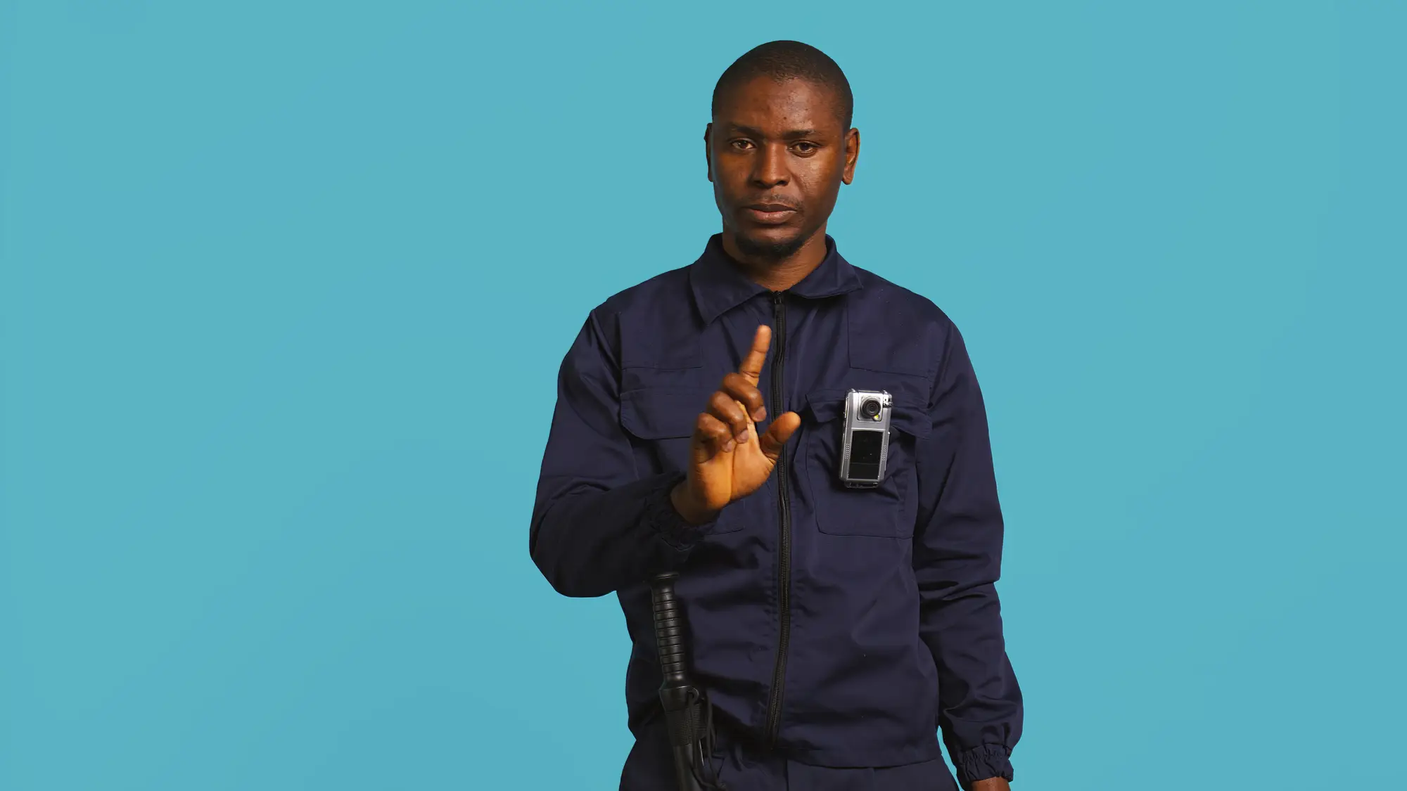 Man in a dark uniform stands against a plain blue background with a body-worn camera on his chest, raising one hand in a stop gesture and looking forward with a serious expression.