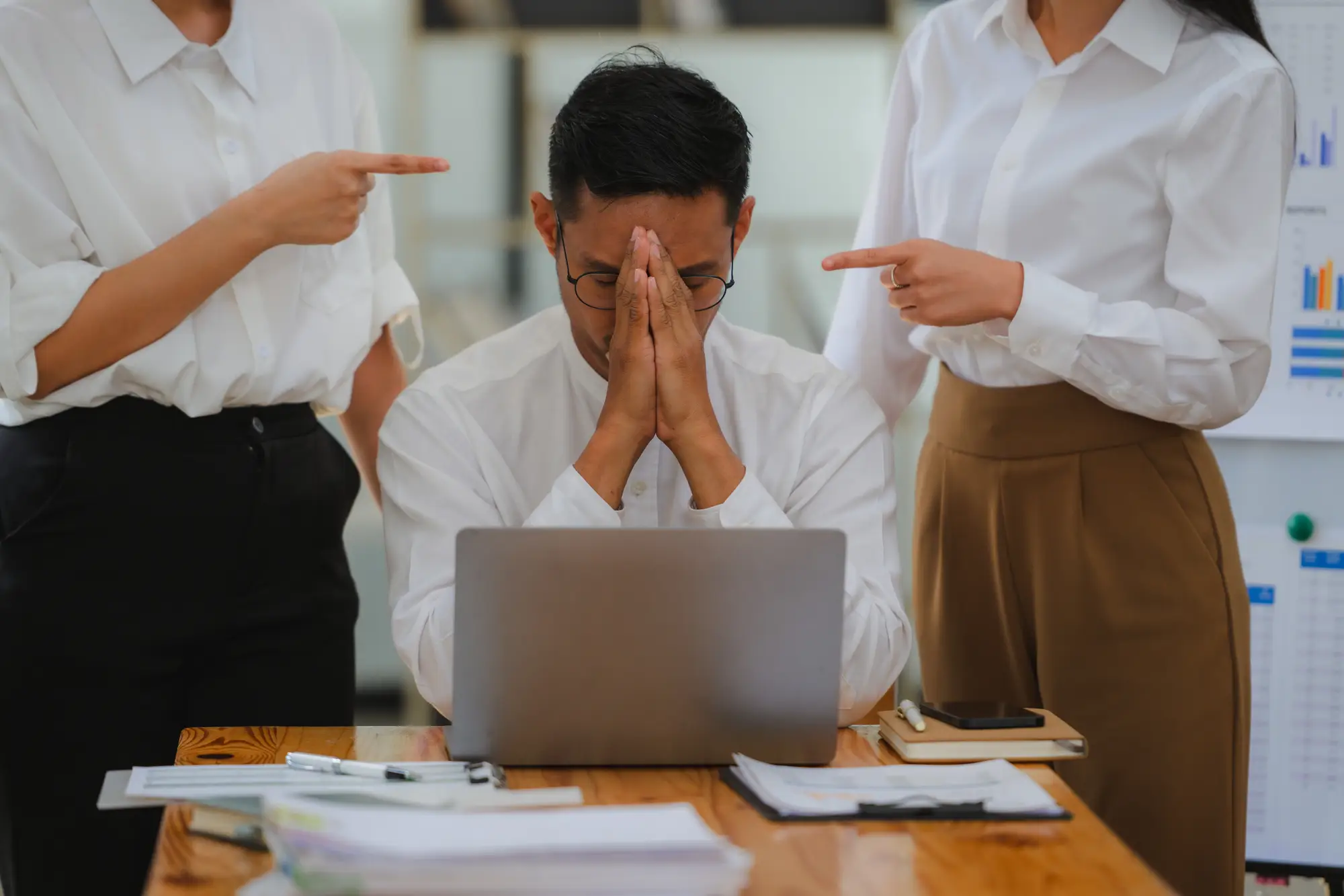 Man in glasses sits stressed at laptop as two coworkers in white point at him in an office
