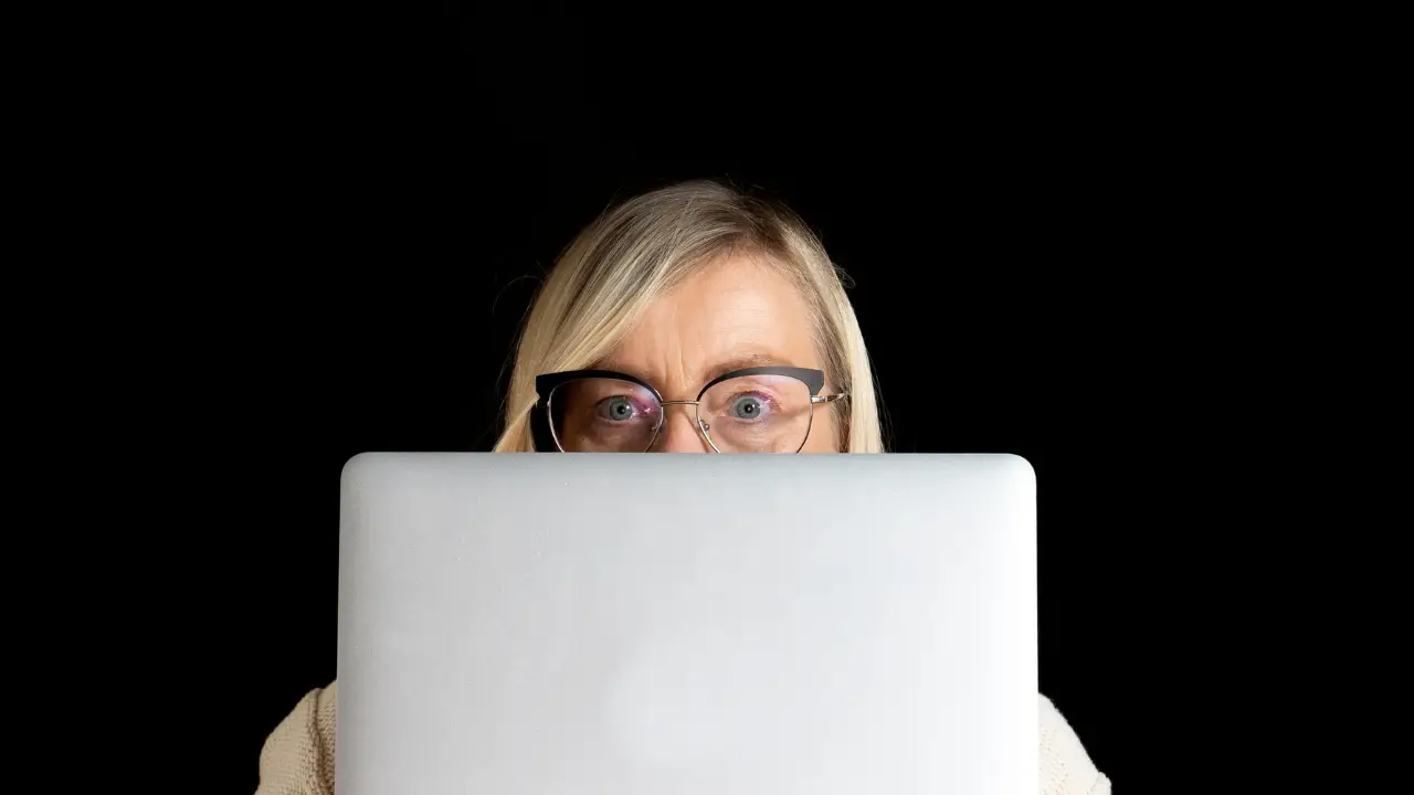 A blonde woman in glasses peers over a laptop with a concerned expression against a dark background