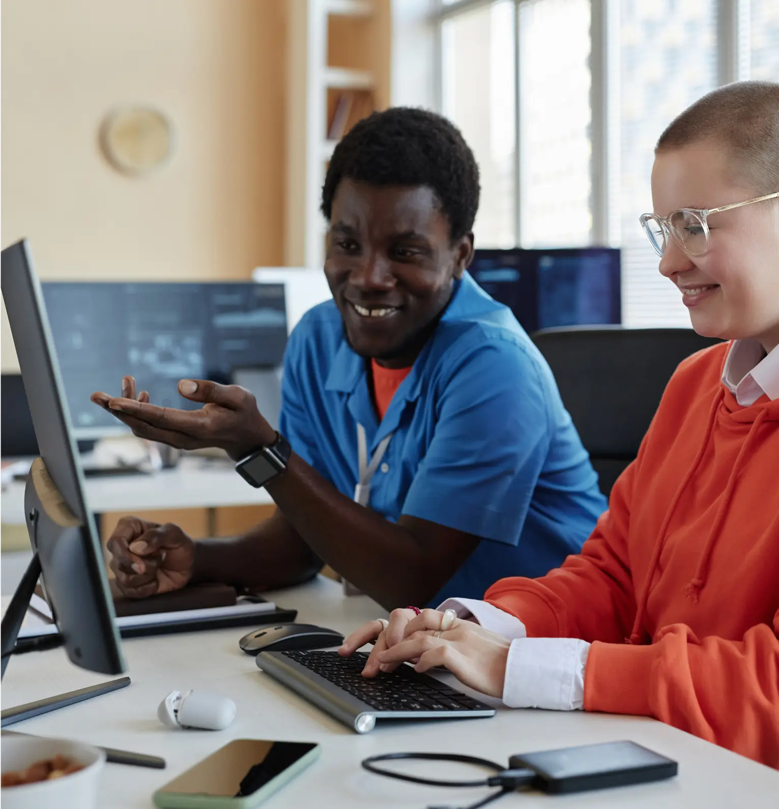 A man in a blue shirt gestures toward the computer screen while a woman in an orange hoodie types on the keyboard. They are smiling as they work together.