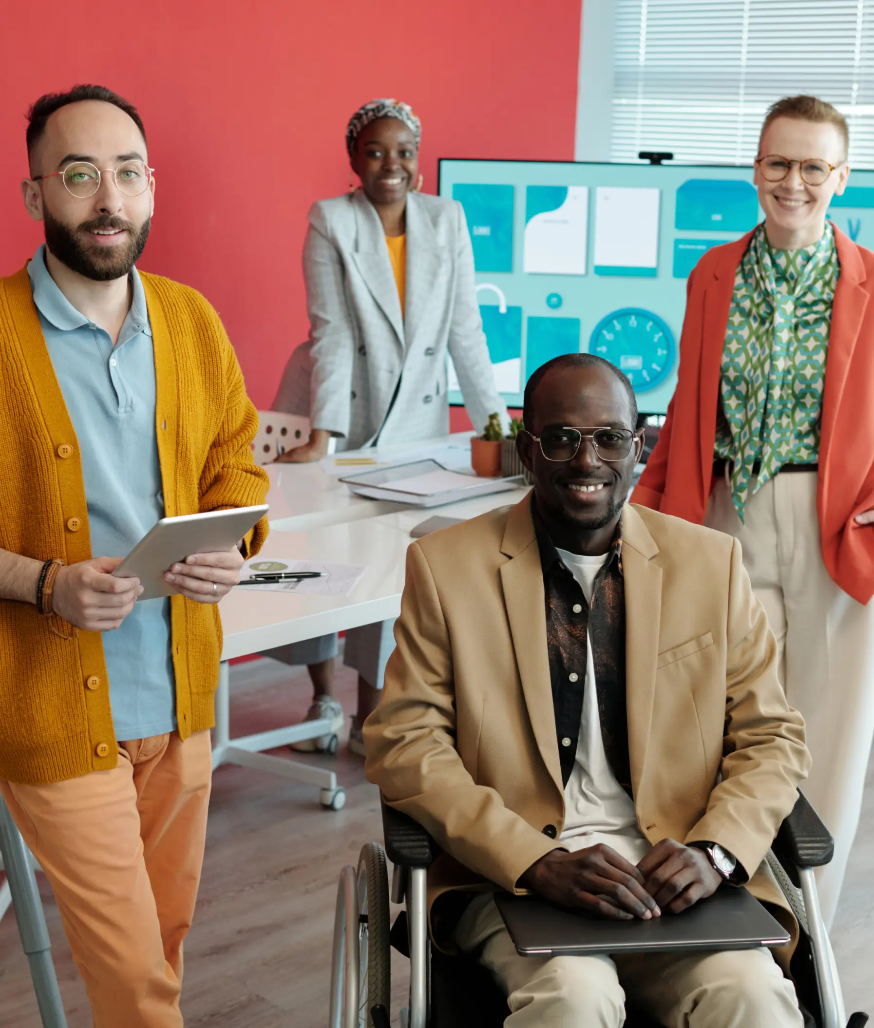 Four professionals gathered in a bright office with a red wall. A man in a mustard cardigan holds a tablet beside a man in a tan blazer seated in a wheelchair. Two women stand behind them, one in a gray suit and one in an orange blazer with a green scarf.