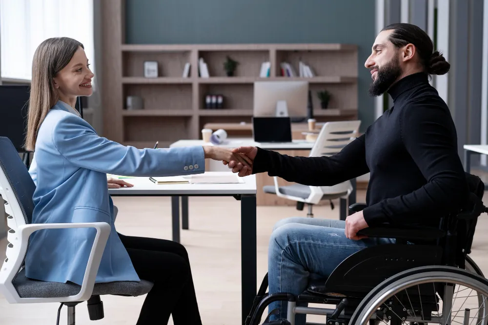 Woman in blue blazer shaking hands with bearded man in wheelchair at desk